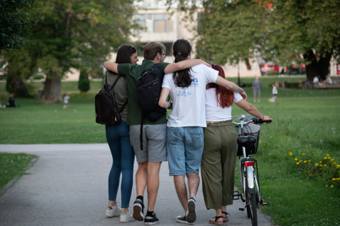 Four young people walking arm in arm along a park path, one pushing a bicycle.